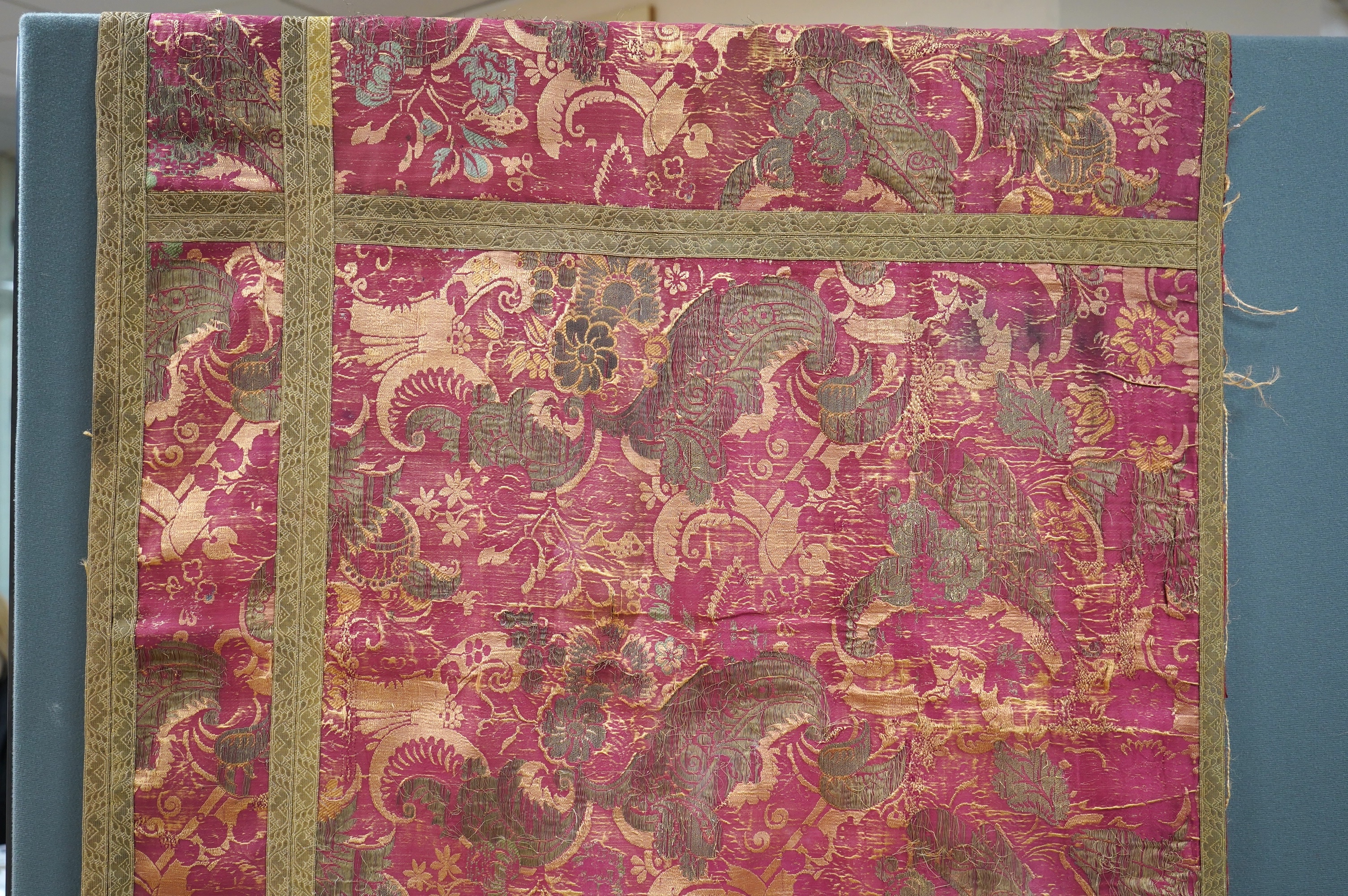 A fine 17th century Italian crimson silk brocade wall hanging, originally woven with gold threads and polychrome silks in a large ornate floral pattern, possibly part of a larger hanging, the front now edged with later n